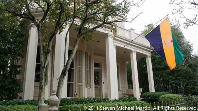 A green, gold, and purple Mardi Gras flag hangs beside a white columned porch framed by trees and shrubs.