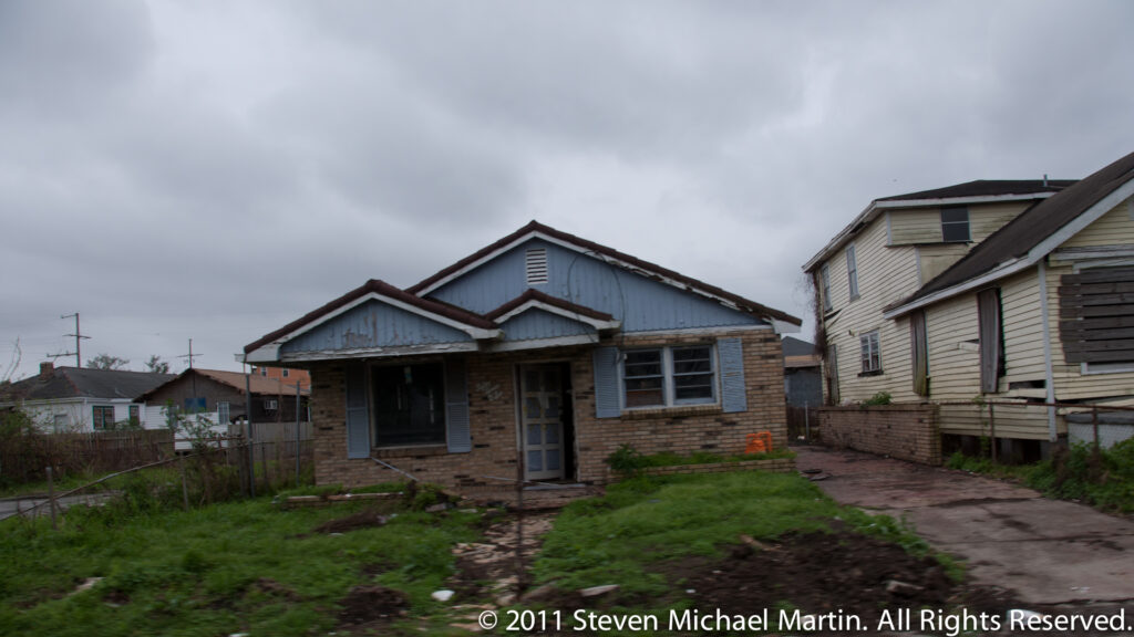 A worn blue-gray house with a shallow porch stands beside closely spaced homes and a patchy muddy yard.