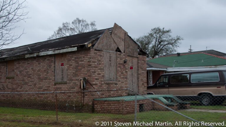 A brick residence with boarded windows stands behind a chain-link fence beside a parked van.