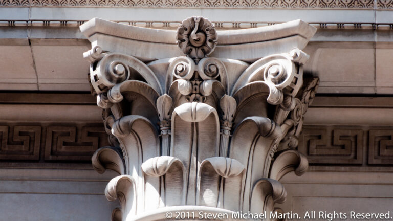 A projecting stone ornament combines a central mask-like face, curled foliage, and acanthus leaves above a Greek-key band.