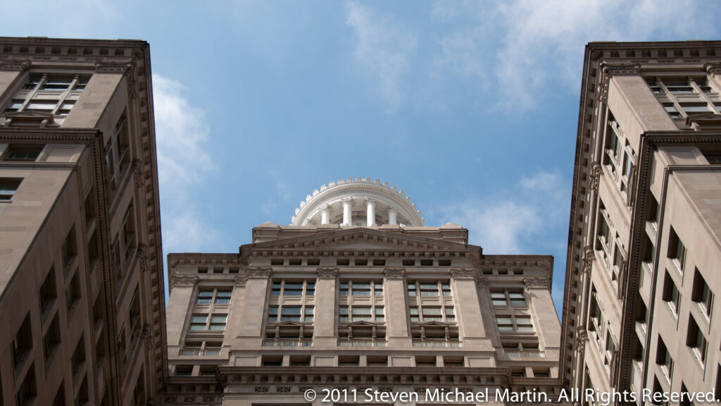 A domed stone tower rises between neighboring office buildings in an upward view from the street.