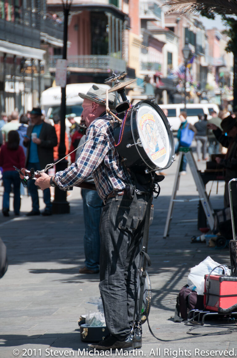 A man in a brimmed hat carries a large drum through a busy pedestrian area.