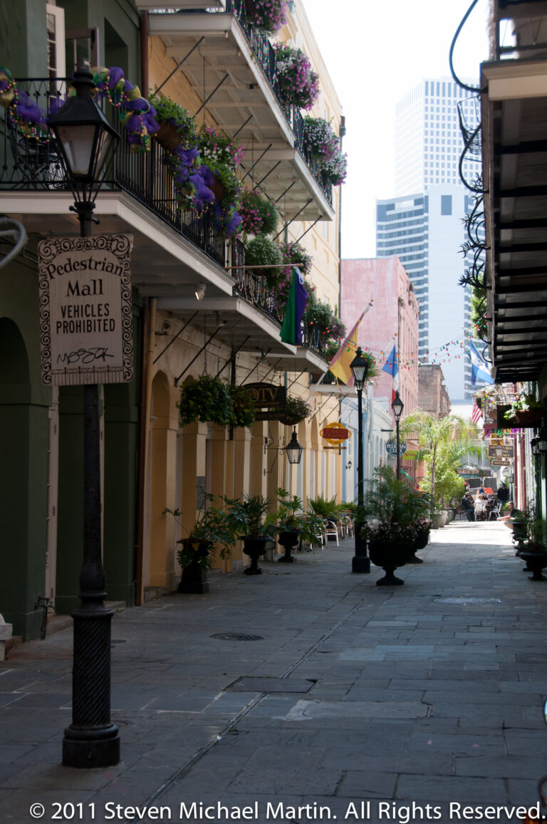 A pedestrian lane with balconies and planters leads toward a distant high-rise; a sign prohibits vehicles.