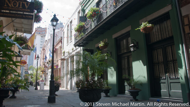 A shaded lane recedes between iron balconies, green doors, potted palms, and a black streetlamp.