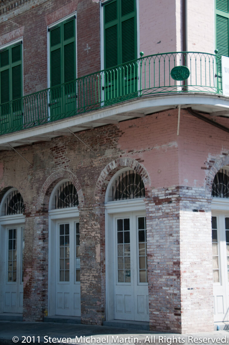 Three arched openings in a brick façade sit below a green iron gallery and shuttered windows.