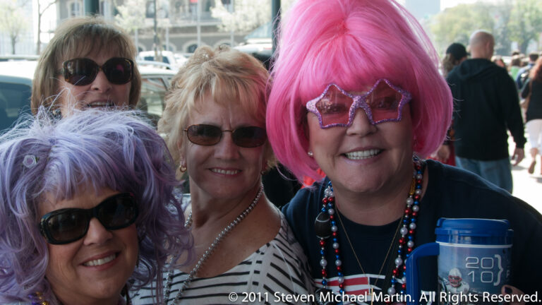 Three smiling women wear colorful wigs, novelty glasses, and bead necklaces among a street crowd.