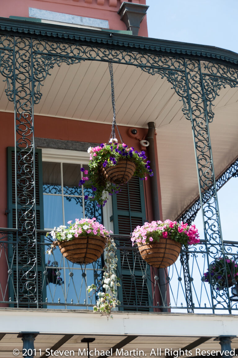 Flower-filled hanging baskets line a wrought-iron balcony above green shutters on a pastel façade.