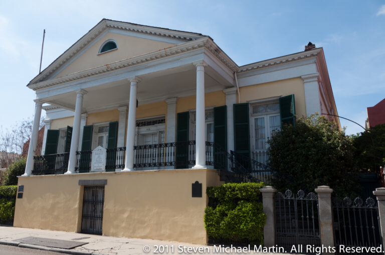 A pale residence with a columned portico, pediment, green shutters, and iron balcony.