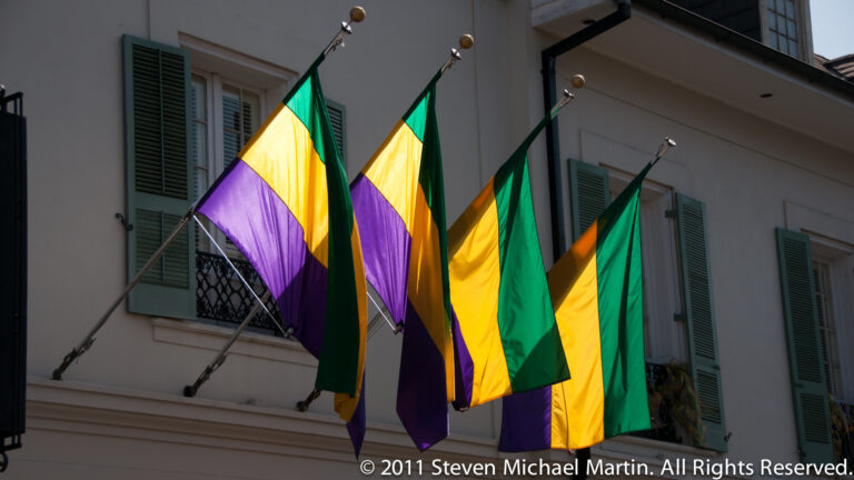 Purple, green, and gold flags hang from a pale building with green shutters and an iron balcony.