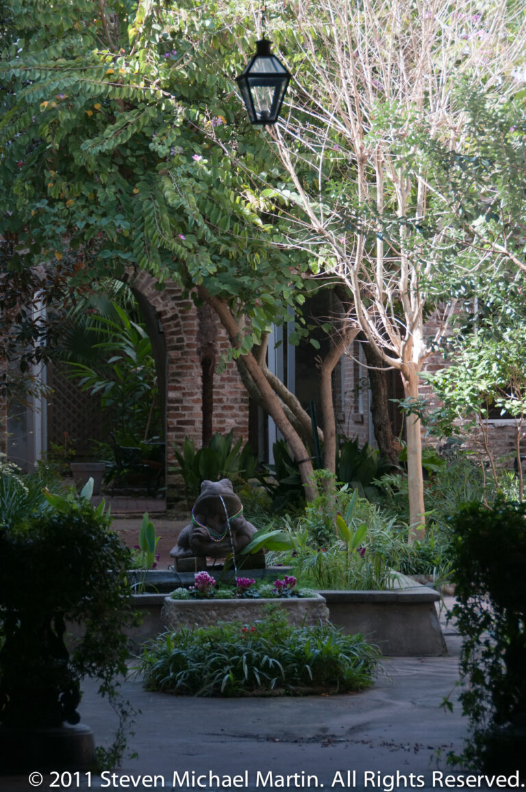 A leafy courtyard garden with a small fountain sits before a shaded brick arcade.