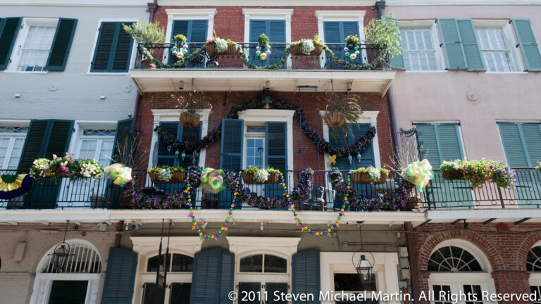 Pastel row buildings display Mardi Gras beads, flower baskets, and colorful balcony decorations.