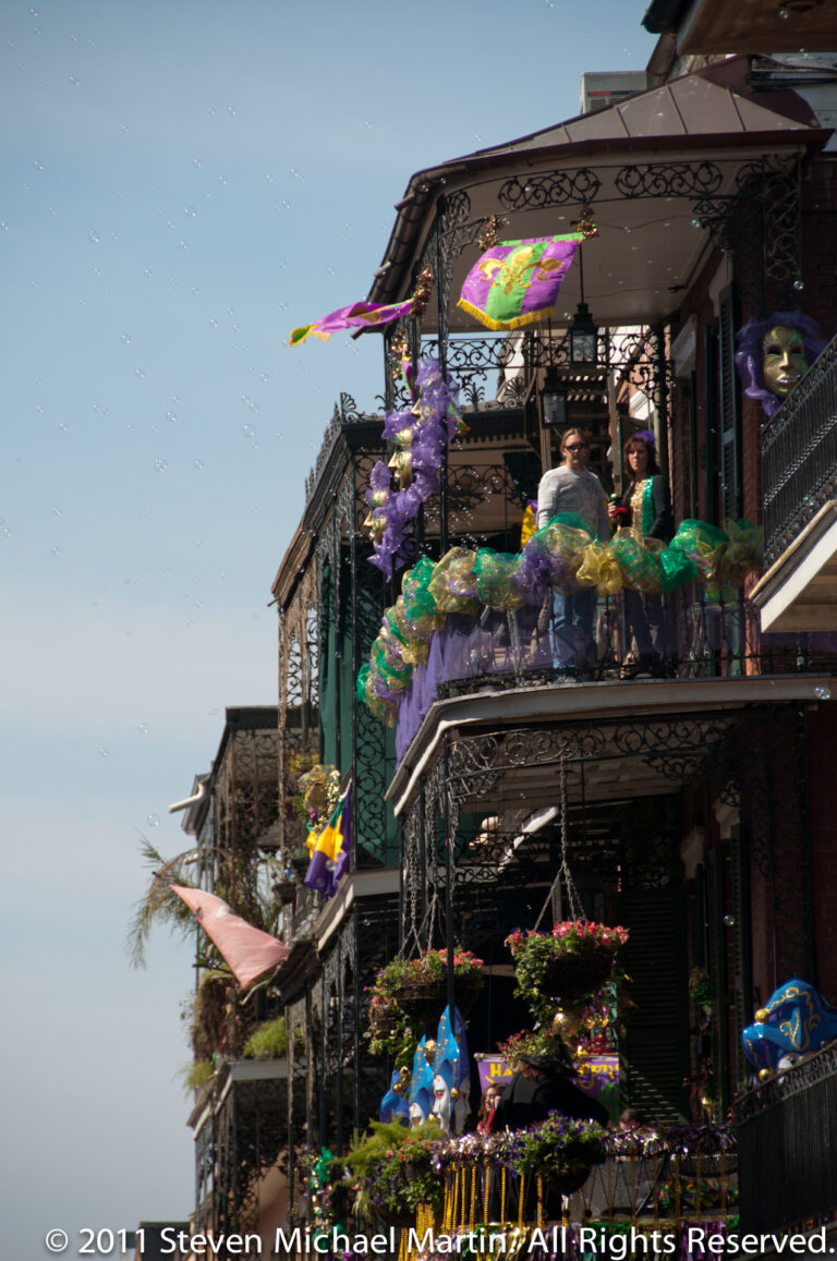 Mardi Gras beads, streamers, and flags decorate stacked iron balconies on a corner building.