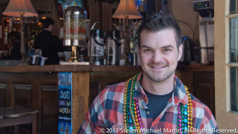 A smiling man in a plaid shirt and bead necklaces poses inside a bar.