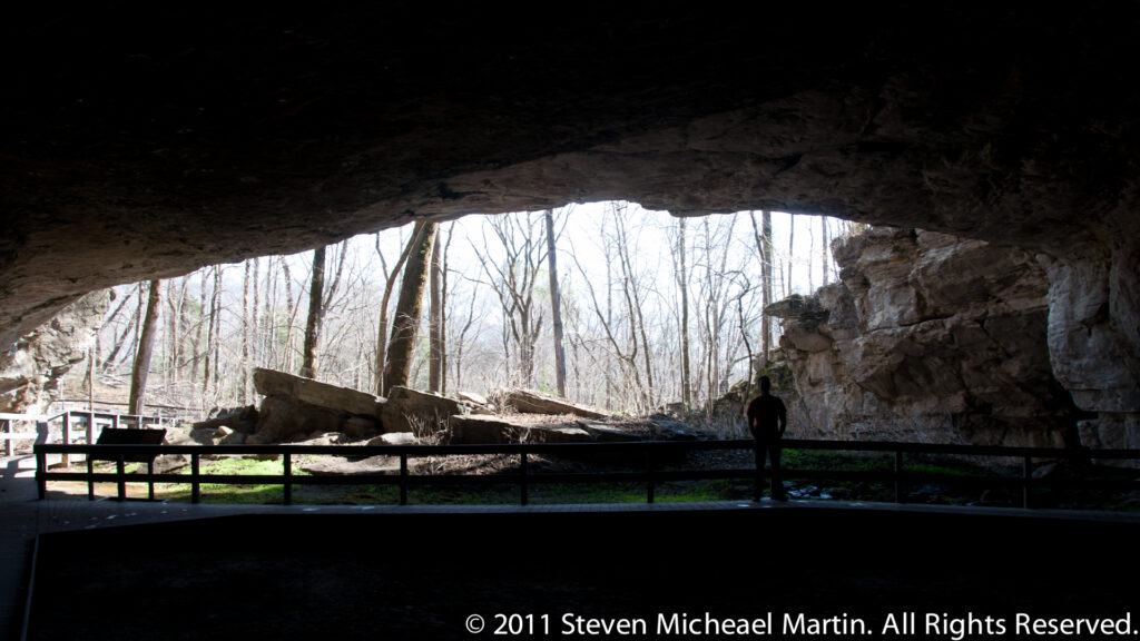 A broad rock shelter frames a sunlit woodland entrance, with a visitor standing behind a low barrier.
