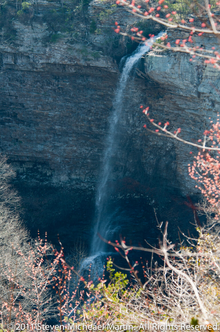 Water drops over a low rock lip into a dark pool, with leaves floating near the edges.