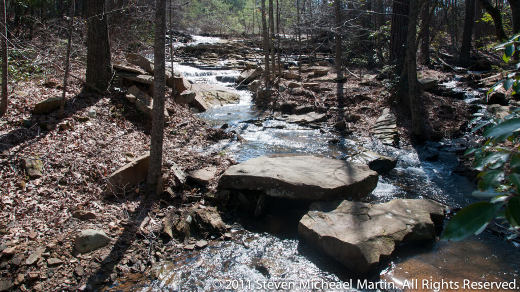 A shallow wooded creek flows around flat rock slabs beneath bare trees.