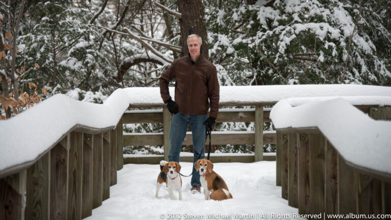 A man stands with two leashed beagles on a snow-covered footbridge surrounded by trees.