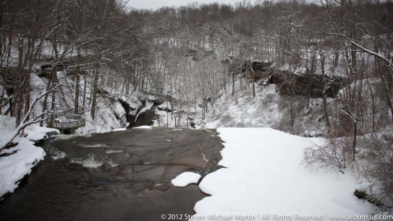 A creek curves through a snowy rocky ravine, with a small footbridge on the left bank.