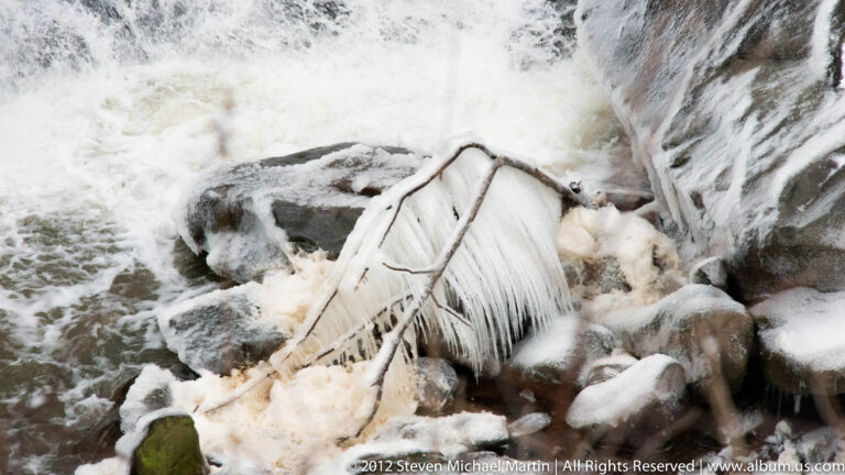 Water rushes past ice-coated rocks and a frozen branch in a snowy ravine.
