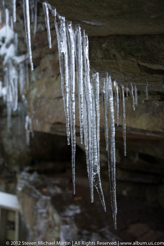 Clear icicles descend from a dark sandstone ledge, with blurred rock behind them.
