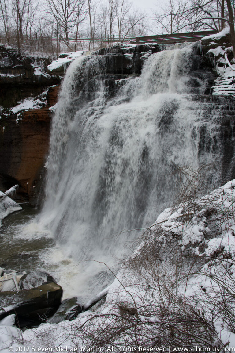 A shallow cascade spills over a dark rock shelf between snowy banks and bare branches.