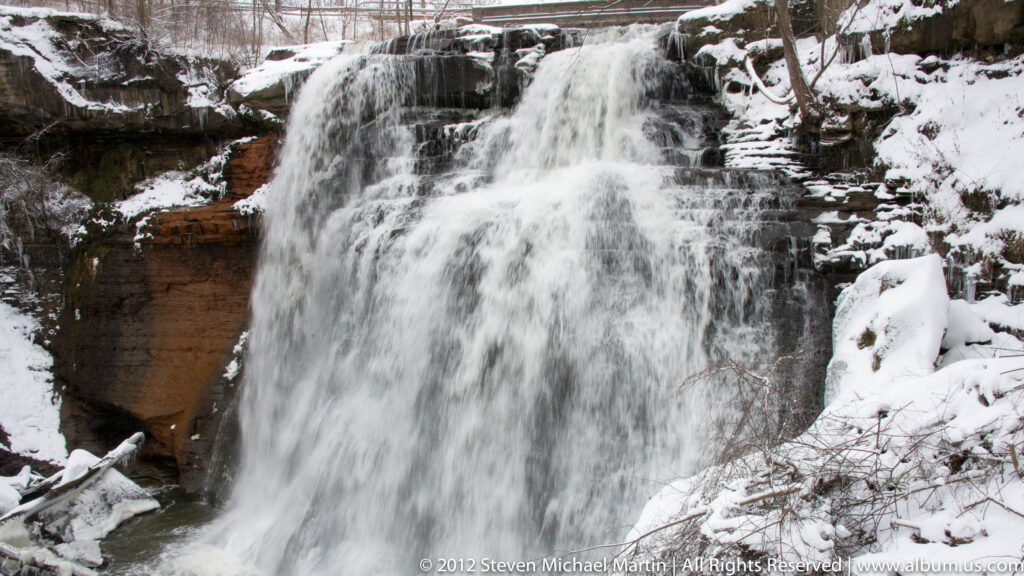 A wide waterfall descends over layered rock, edged by icicles and snow-covered trees.