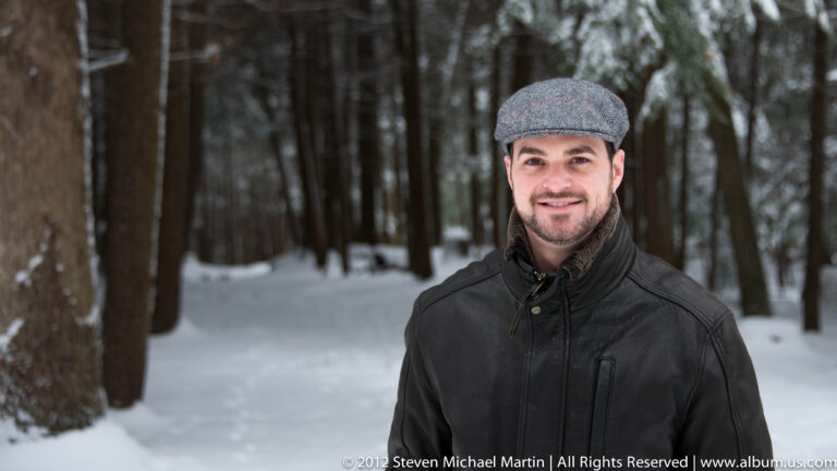 A smiling man in a gray cap and dark jacket stands among snowy trees.
