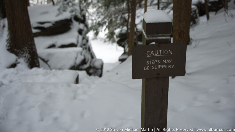A brown caution sign warns that steps may be slippery beside a snow-covered woodland stairway.
