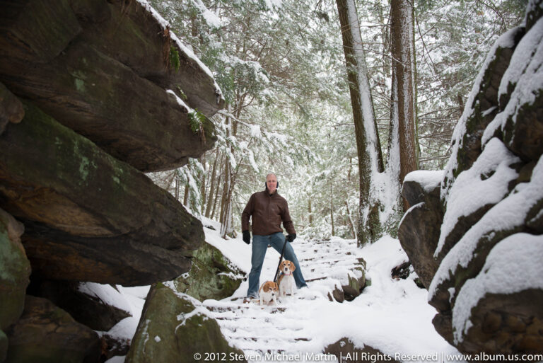 A man and two beagles descend snowy stone steps between large sandstone boulders.