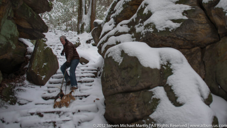A man follows two leashed beagles down snowy steps between close-set boulders.