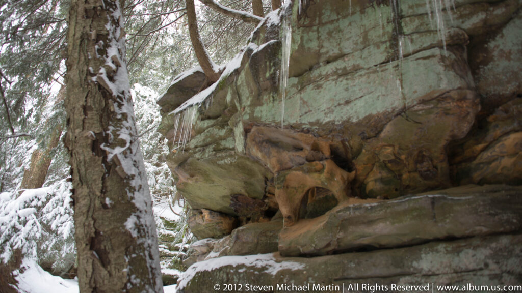 A deer rests on a ledge beneath a sandstone overhang, with snow on the surrounding rocks.