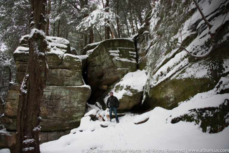 A visitor and two beagles stand beside a huge boulder with a dark crevice behind them.