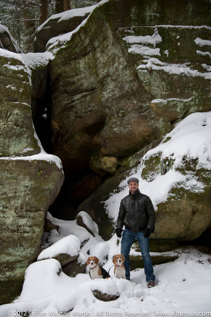 A man stands behind two beagles beneath a broad sandstone overhang with a dark recess.