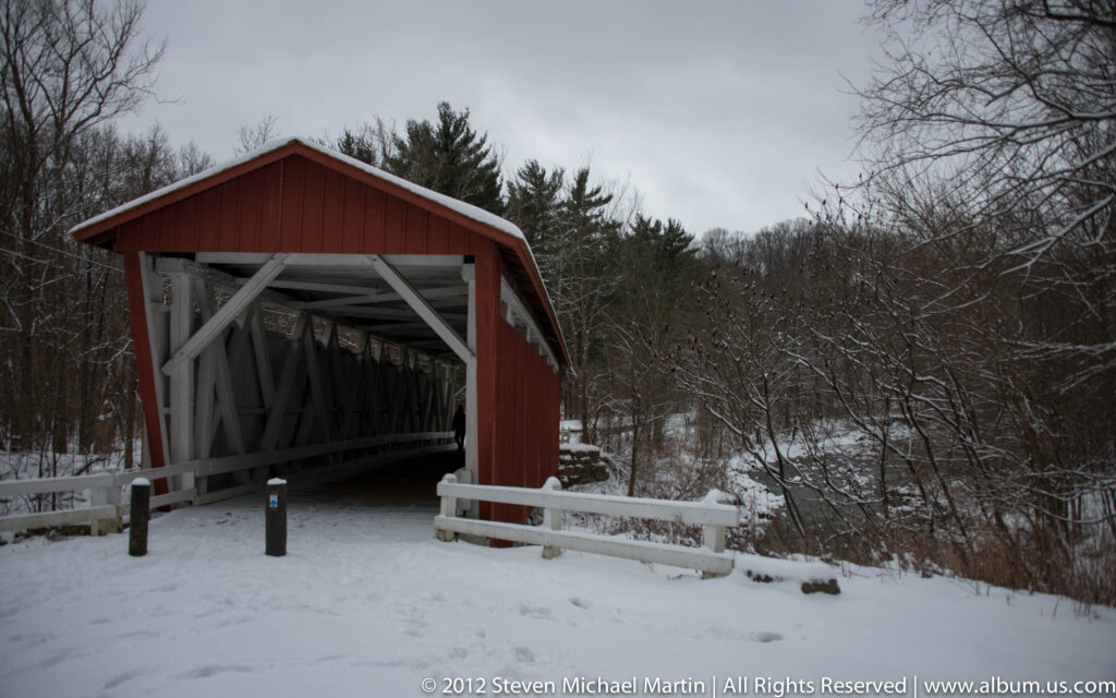 A red covered bridge with white trusses stands beside a snowy wooded valley.