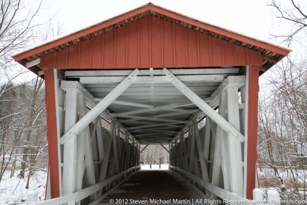 White timber trusses and braces form a long interior passage inside a red covered bridge.
