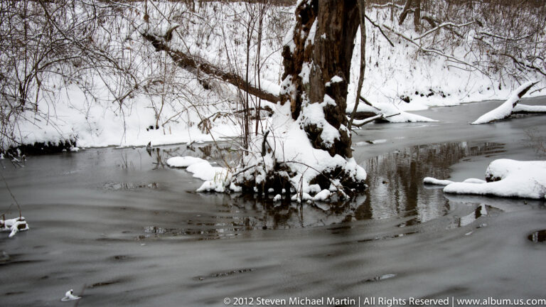 A partly frozen creek curves past a snow-covered tree and rocky banks in winter woods.