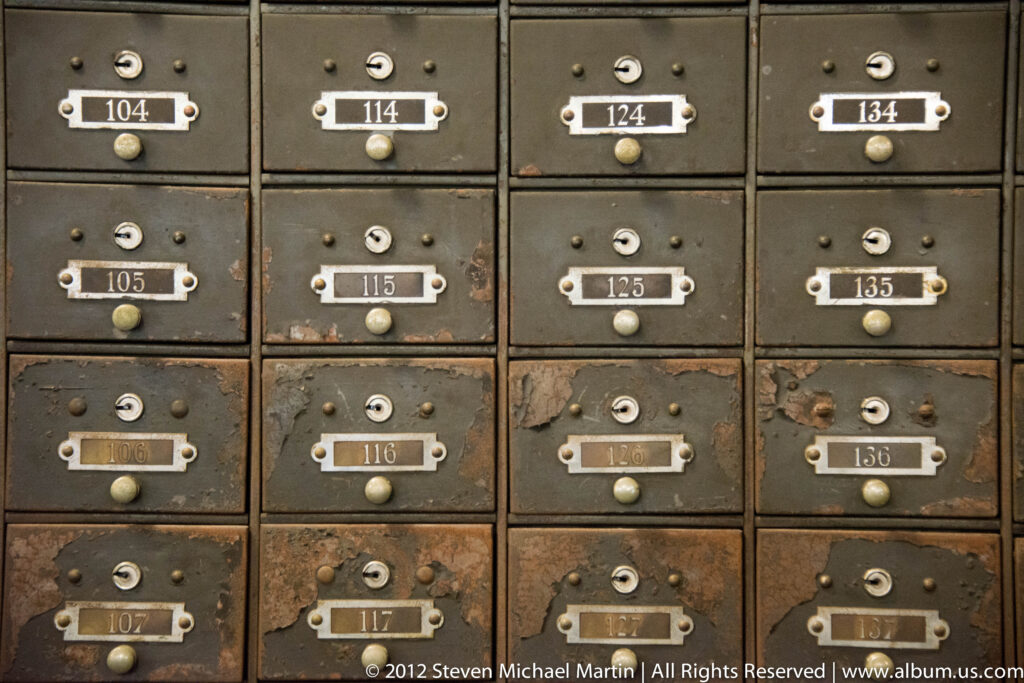 A worn metal cabinet has rows of small numbered compartments, keyholes, knobs, and nameplates.