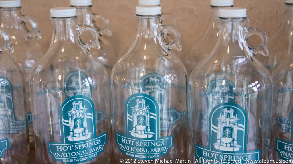 Rows of clear bottles carry turquoise Hot Springs National Park thermal-water labels with a bathhouse illustration.