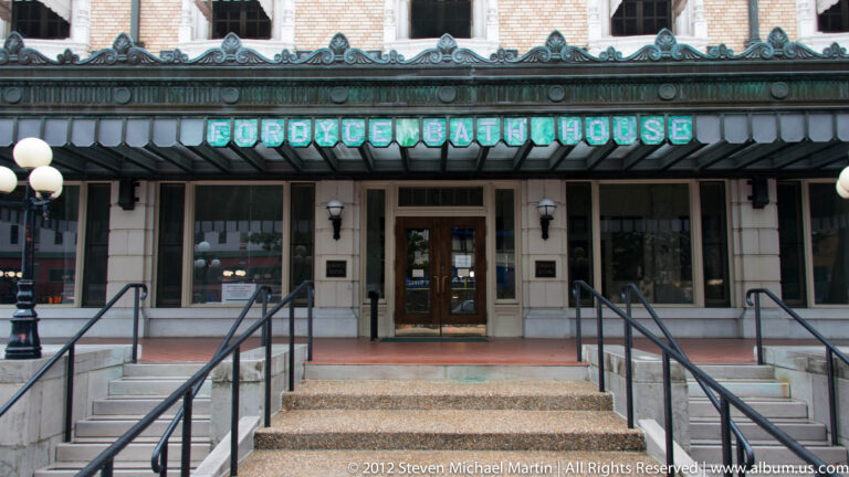 The Fordyce Bathhouse sign sits above glass doors reached by broad steps and paired metal railings.