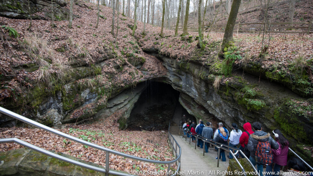 Visitors descend a metal-railed stairway toward a wide cave mouth beneath a wooded limestone ledge.