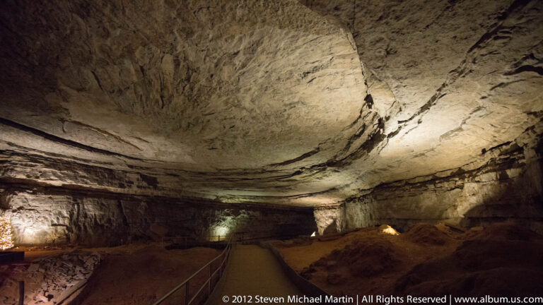 A lit path crosses a broad cave chamber beneath a low layered ceiling of limestone.