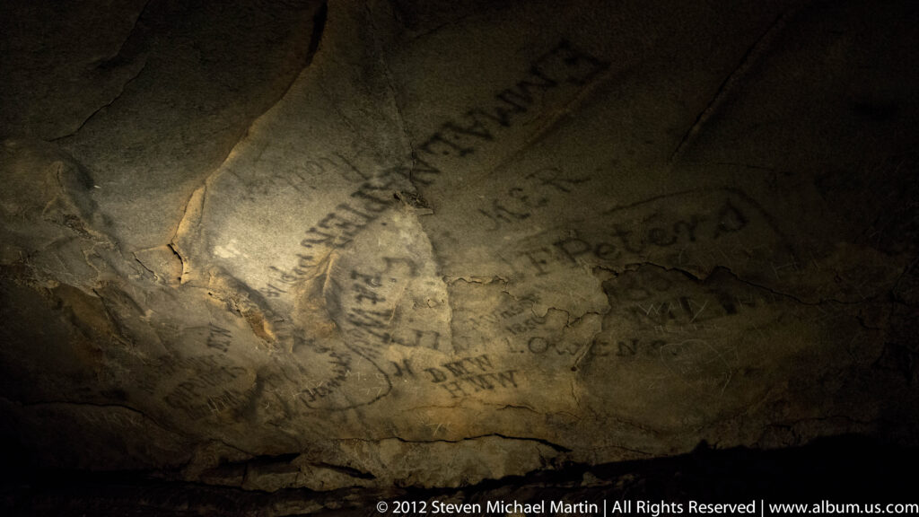 A beam of light reveals numerous handwritten names and marks across a pale cave wall.