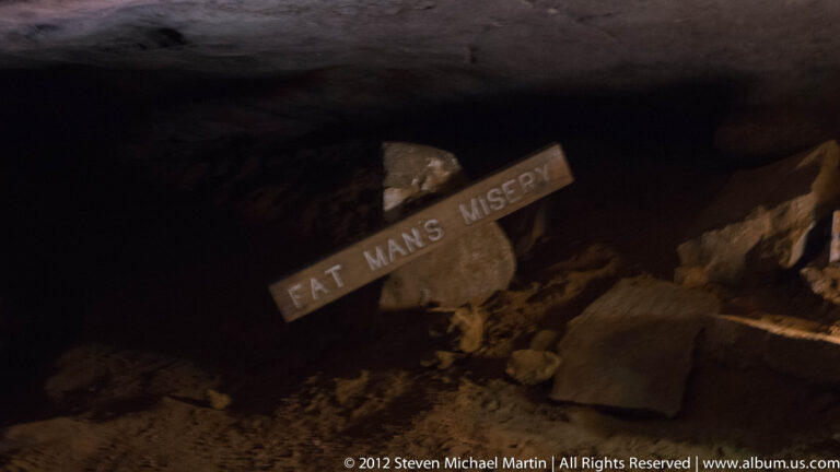A sign reading “Fat Man’s Misery” stands beside a narrow, low passage in a dark cave.