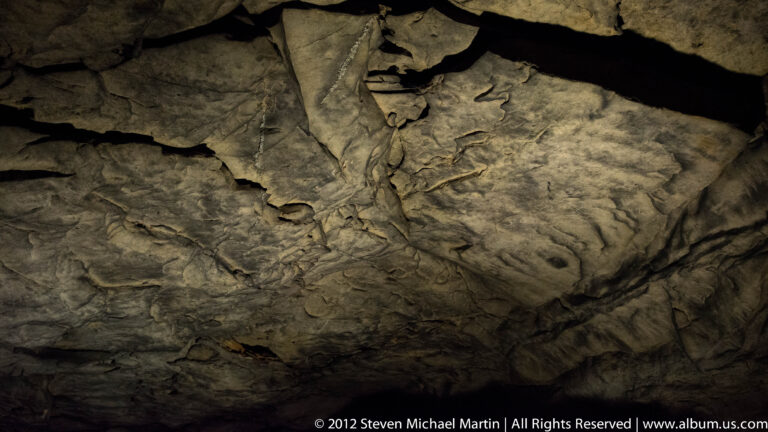 Direct light reveals fractured plates and dark fissures across a rugged limestone cave ceiling.