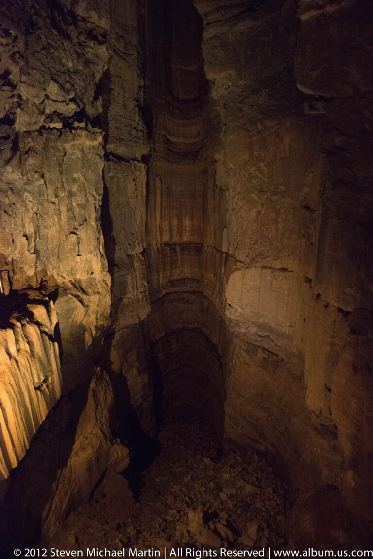 A dark recess lies beneath broad layered rock ledges in a cave chamber lit by warm lamps.