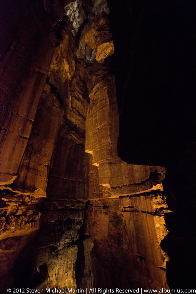 An amber-lit column of layered limestone rises between a sloping wall and a dark cave void.