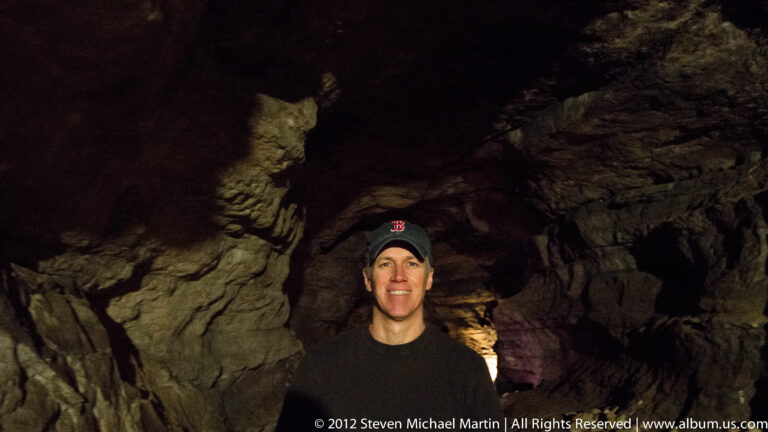 A smiling visitor wearing a baseball cap stands before rough rock walls in a dim cave.