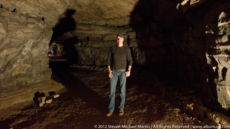 A man stands in a broad cave chamber beneath a high rock ceiling, with another visitor in the distance.
