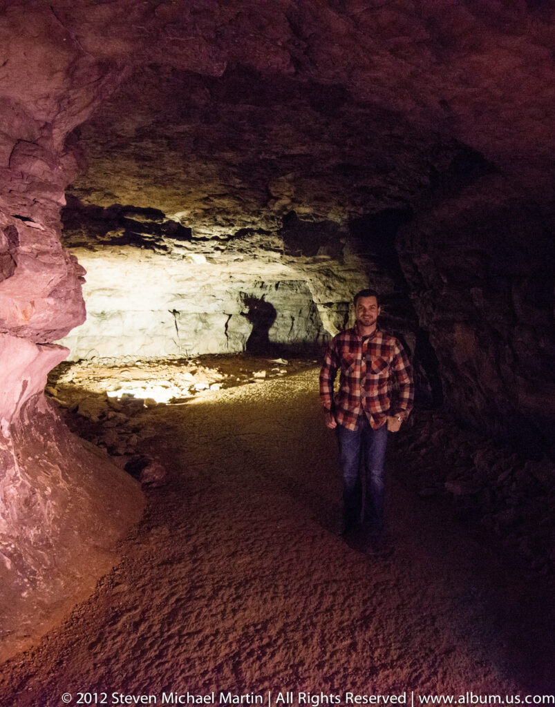 A visitor in a plaid shirt stands beside a reflective wet floor beneath a brightly lit cave wall.