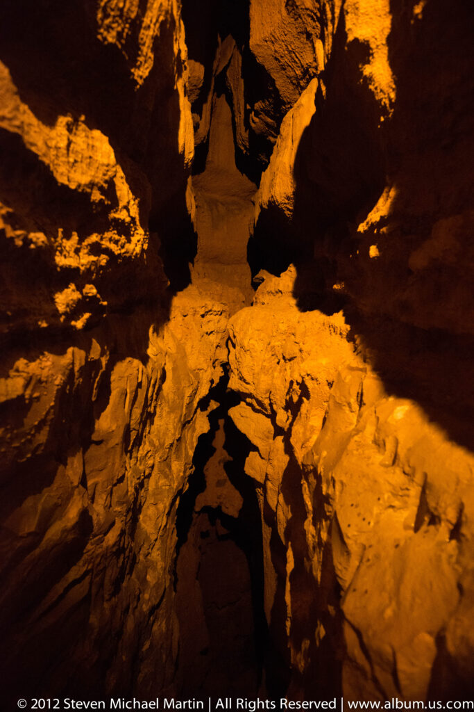 Amber light illuminates jagged rock walls around a narrow dark passage through the cave.